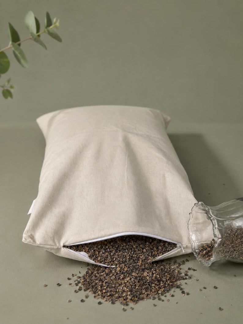 May include: A rectangular beige fabric pillow with a white zipper closure partially open at the bottom. Dark brown buckwheat hulls are spilling out from the opening onto the surface, and a clear glass vessel is positioned next to the pillow.
