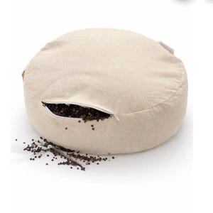 Op de afbeelding: Een ronde, beige zitzak met een ritssluiting, die een donkerbruine vulling onthult. Een deel van de vulling is op het witte oppervlak gemorst. De zitzak is gemaakt van een textuurstof.