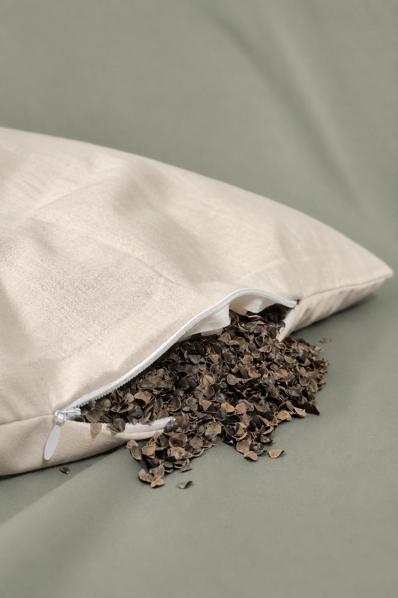 May include: A rectangular off-white fabric pillowcase with a white zipper closure. The pillowcase is partially open, revealing a filling of dark brown, triangular buckwheat hulls that are spilling out from the opening onto the surface below.