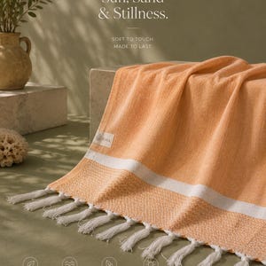 May include: An orange beach towel with white stripes and white fringe. The towel is draped over a stone block. Text on the towel reads "Made for Sun, Sand & Stillness." Additional text includes "Lightweight & Breathable" and "Quick Dry & Absorbent."