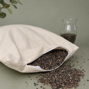 May include: A square, off-white fabric pillow case with a white zipper closure, partially filled with dark brown buckwheat hulls. Some of the buckwheat hulls have spilled out from the open zipper onto the surface in front of the pillow.