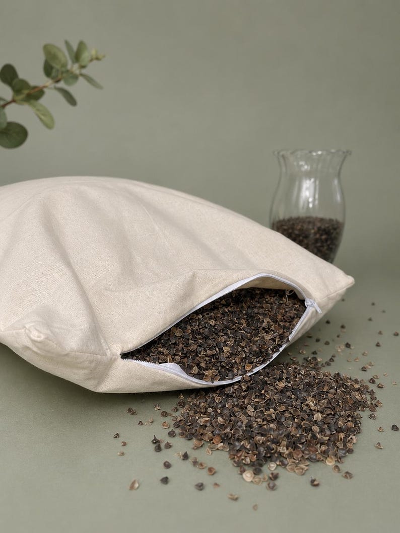 May include: A square, off-white fabric pillow case with a white zipper closure, partially filled with dark brown buckwheat hulls. Some of the buckwheat hulls have spilled out from the open zipper onto the surface in front of the pillow.