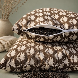 May include: Two brown throw pillows with a white floral pattern are stacked. One pillow is partially unzipped, revealing a dark filling. A small pile of the same dark filling sits in front of the pillows. A vase with dried plants is in the background.