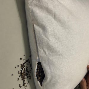 May include: A square, off-white linen pillow cover featuring a side zipper closure. The zipper is partially open, revealing a dark brown buckwheat hull filling inside. A small pile of loose buckwheat hulls is visible next to the pillow.
