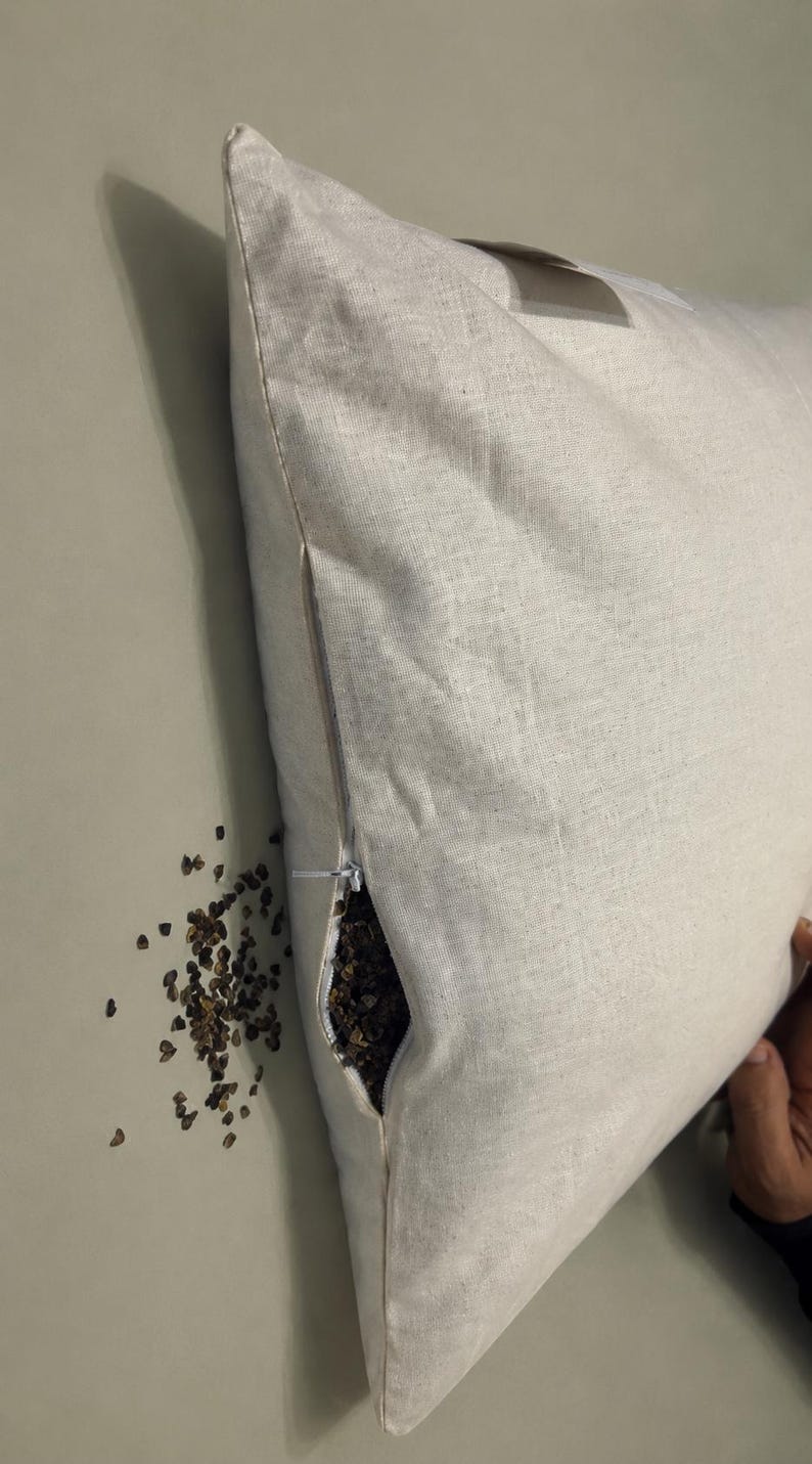 May include: A square, off-white linen pillow cover featuring a side zipper closure. The zipper is partially open, revealing a dark brown buckwheat hull filling inside. A small pile of loose buckwheat hulls is visible next to the pillow.