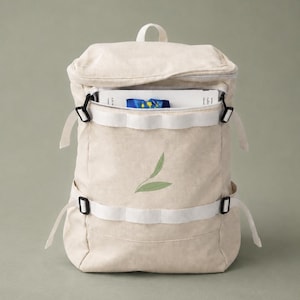 Può includere: Zaino in tela beige con cinghie bianche e fibbie nere. Lo zaino ha una chiusura a patta e una tasca frontale. Un disegno a foglia verde è stampato sul davanti. La borsa è progettata per trasportare libri e altri oggetti.