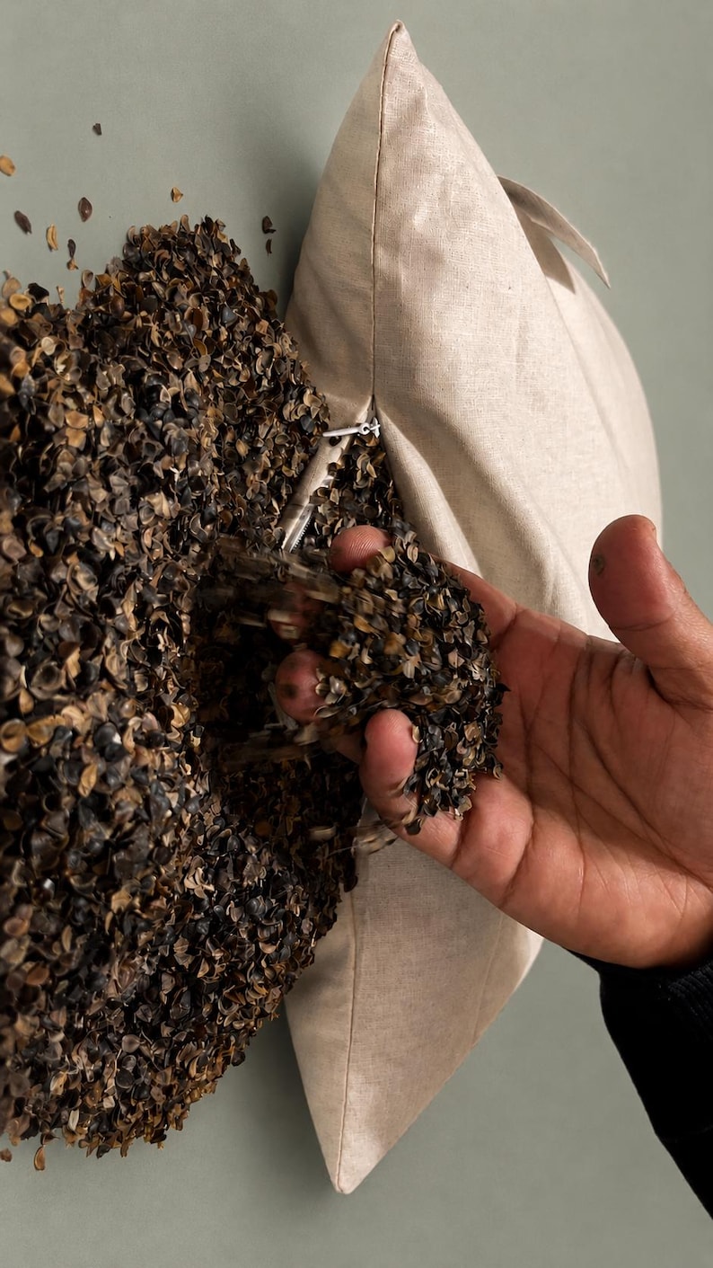 May include: A beige linen pillowcase partially filled with dark brown buckwheat hulls. The pillowcase features a visible zipper closure along the side, and the dark hulls are spilling out from the opening, showing their textured, irregular shape.