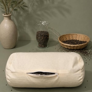 May include: A cylindrical off-white bolster pillow made of plain fabric with a central horizontal zipper opening. The zipper is partially open, revealing a dark, granular filling inside the pillow. The pillow has a simple, unadorned design.