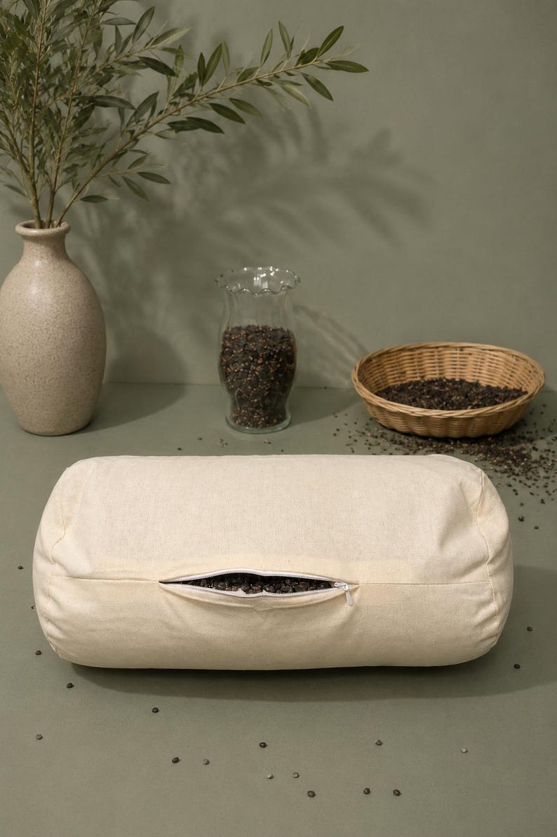 May include: A cylindrical off-white bolster pillow made of plain fabric with a central horizontal zipper opening. The zipper is partially open, revealing a dark, granular filling inside the pillow. The pillow has a simple, unadorned design.