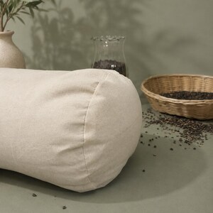 May include: A beige linen bolster cushion, cylindrical in shape. It rests on a sage green surface, with a small woven basket and a glass jar in the background. The scene is illuminated by natural light.
