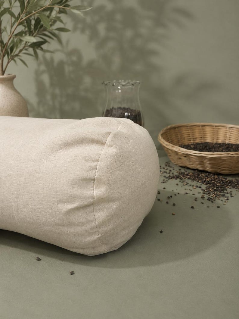 May include: A beige linen bolster cushion, cylindrical in shape. It rests on a sage green surface, with a small woven basket and a glass jar in the background. The scene is illuminated by natural light.