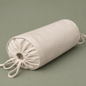 May include: A cylindrical bolster pillow in a neutral beige color. The pillow has a drawstring closure on both ends and is made of a soft, textured fabric. The pillow is approximately 16 inches long.
