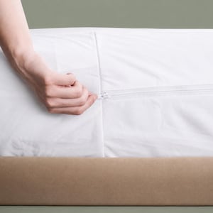 May include: A white mattress cover with a zip closure, being opened. The zip is being pulled by a hand. The cover is on a bed with a tan base.