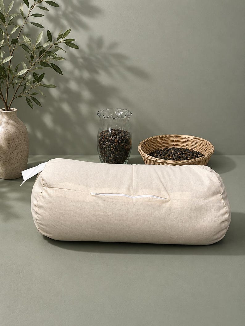 May include: A cylindrical bolster pillow in a solid light beige fabric. The pillow features a horizontal white zipper closure along the front side and a small white fabric tag attached to the left end, presenting a simple and functional design.