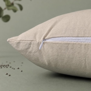 May include: A beige throw pillow cover made of a textured fabric featuring a white hidden zipper closure along the side seam. The pillow cover has a visible weave pattern and is shown from a side angle, highlighting the construction of the seam.