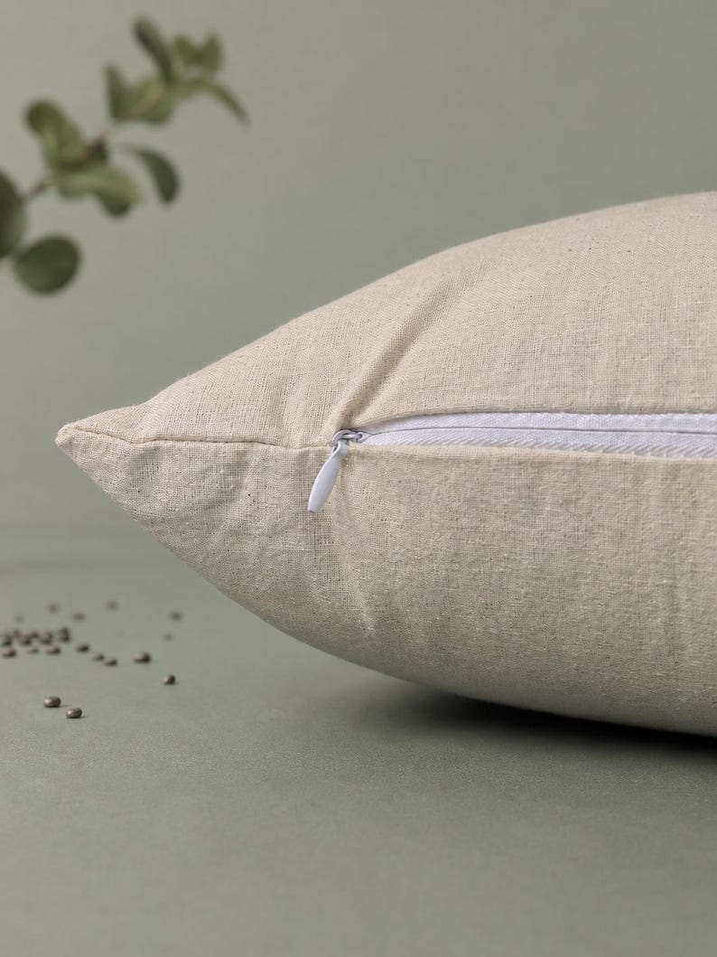 May include: A beige throw pillow cover made of a textured fabric featuring a white hidden zipper closure along the side seam. The pillow cover has a visible weave pattern and is shown from a side angle, highlighting the construction of the seam.