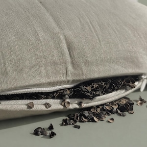 May include: Close-up of a light beige pillow with a partially open zipper revealing dark brown buckwheat hulls. The pillow is made of a textured fabric, and some hulls are scattered around the zipper.