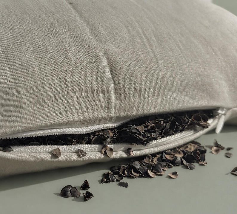 May include: A beige linen pillow cover with a white zipper closure, partially open to reveal a filling of dark brown buckwheat hulls. Some of the buckwheat hulls have spilled out from the opening onto the surface below the pillow.