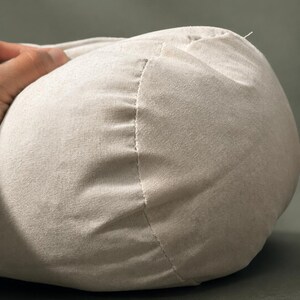 May include: Close-up of a beige, rounded fabric object with a visible seam. The fabric appears to be a soft, woven material. The object is partially held by a hand, suggesting it is a pillow or cushion.