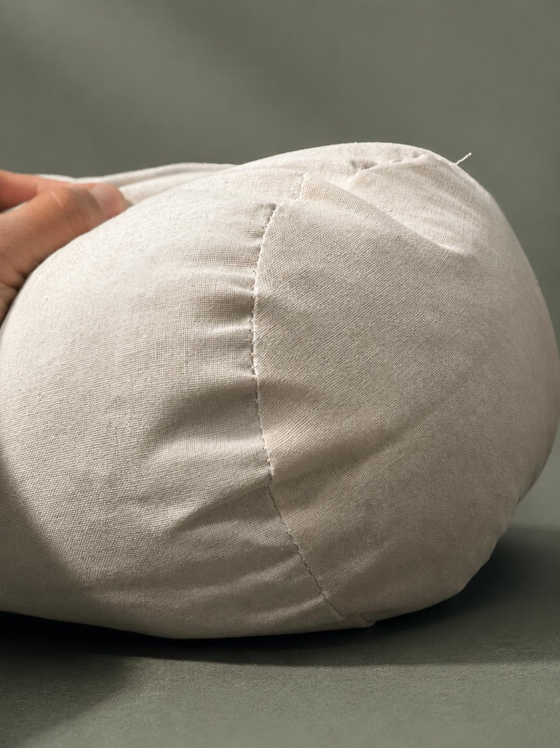 May include: Close-up of a beige, rounded fabric object with a visible seam. The fabric appears to be a soft, woven material. The object is partially held by a hand, suggesting it is a pillow or cushion.