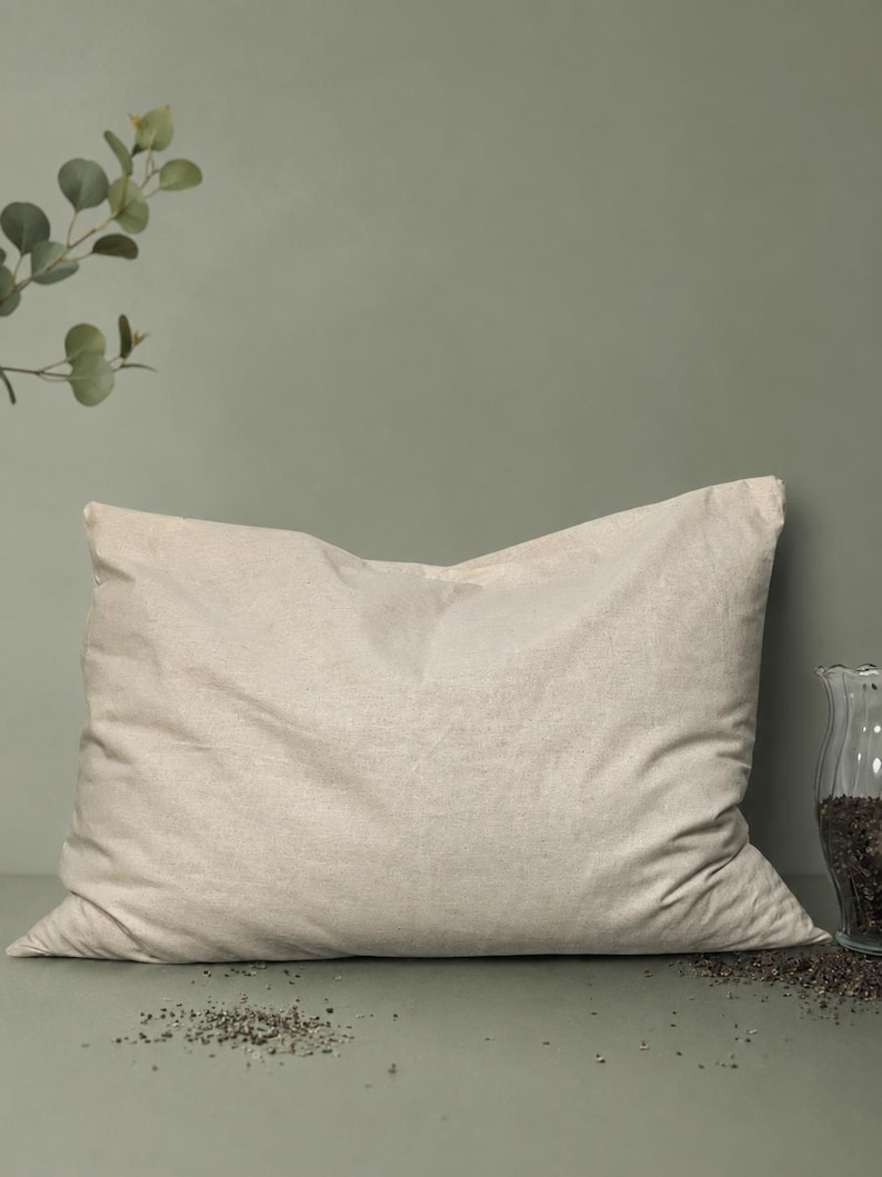 May include: A rectangular pillow with a plain, light beige fabric cover. The pillow is positioned horizontally, featuring a soft, slightly wrinkled texture across its surface. To the right, a clear glass container holds dark, granular contents.
