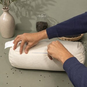 May include: A cream-coloured bolster pillow with a zip closure. The pillow is being unzipped by a person wearing a navy blue long-sleeved shirt. The background includes a vase with greenery and a small glass jar.