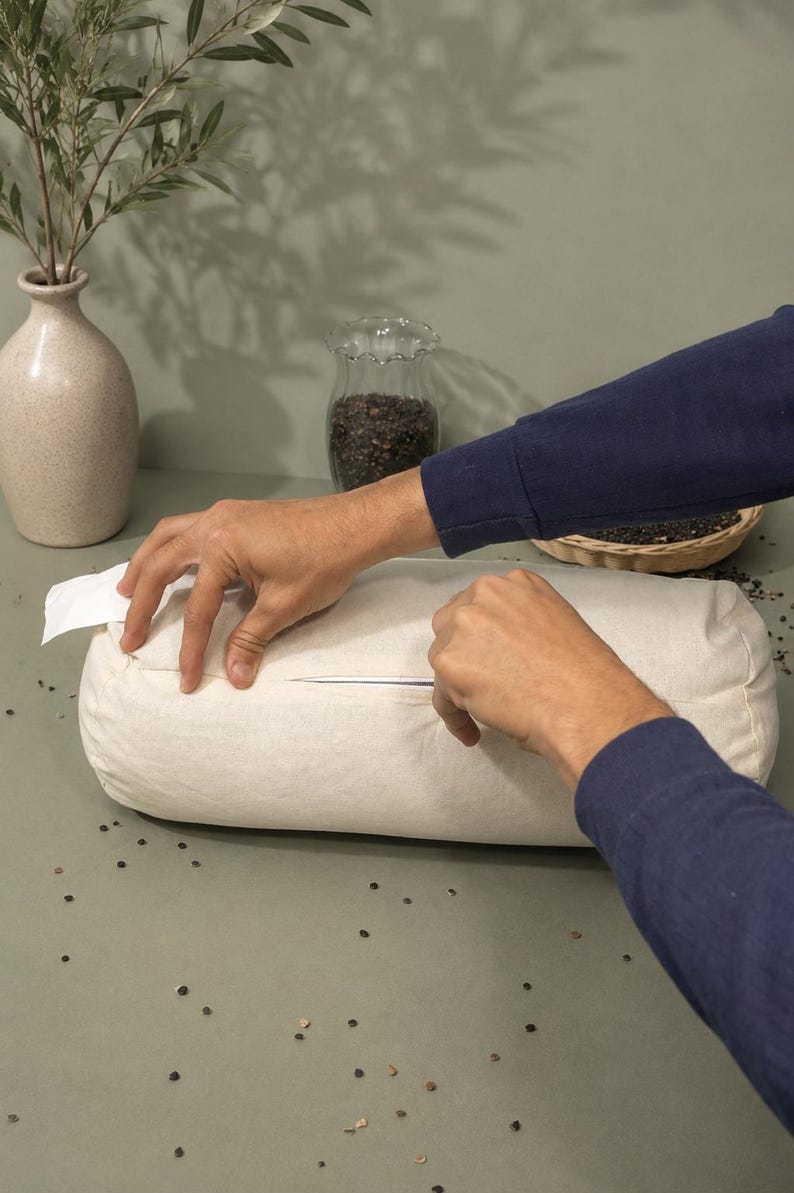 May include: A cream-coloured bolster pillow with a zip closure. The pillow is being unzipped by a person wearing a navy blue long-sleeved shirt. The background includes a vase with greenery and a small glass jar.