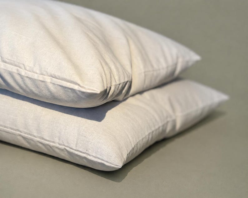 May include: Two rectangular pillows stacked on top of each other, featuring a plain, off-white, textured fabric cover with visible stitching along the edges. The pillows appear soft and filled, with a simple, clean design and a neutral color tone.