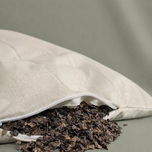 May include: A beige linen pillowcase with a white zipper closure, partially open to reveal a filling of dark brown buckwheat hulls spilling out. The fabric has a visible woven texture, and the pillow is filled with loose, organic grain husks.