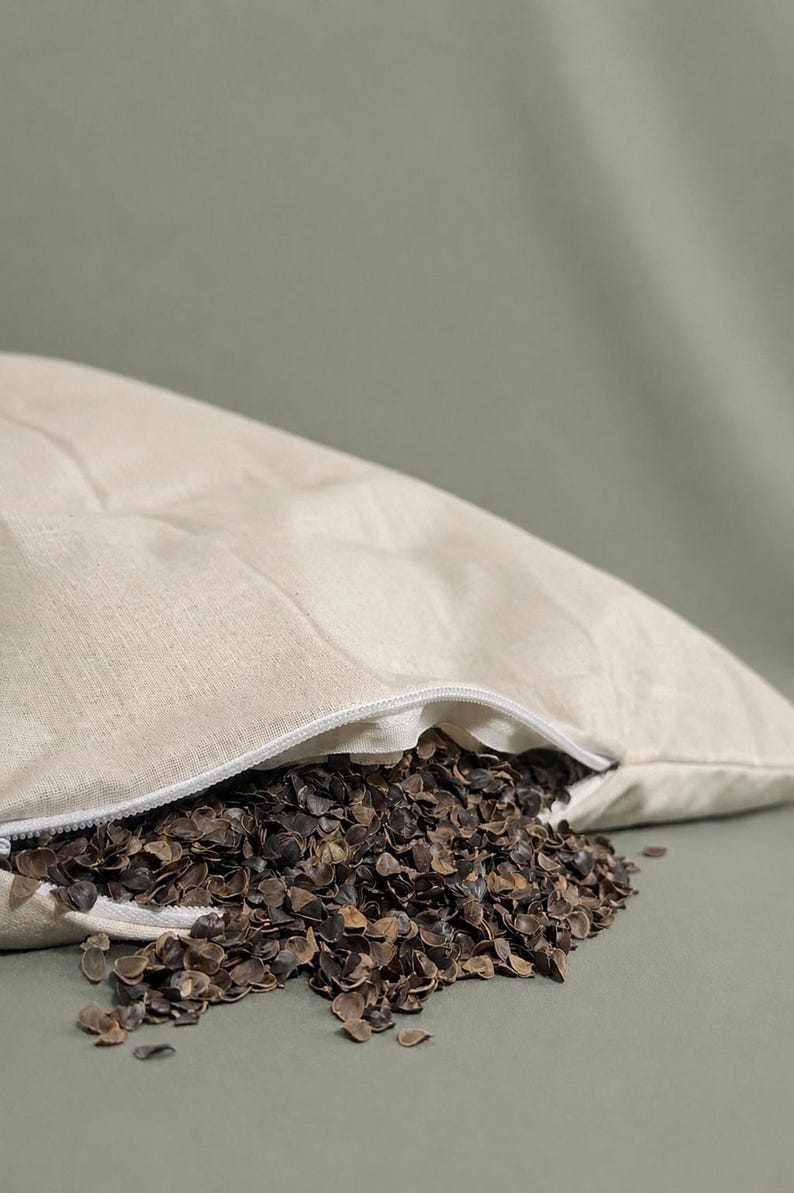 May include: A beige linen pillowcase with a white zipper closure, partially open to reveal a filling of dark brown buckwheat hulls spilling out. The fabric has a visible woven texture, and the pillow is filled with loose, organic grain husks.