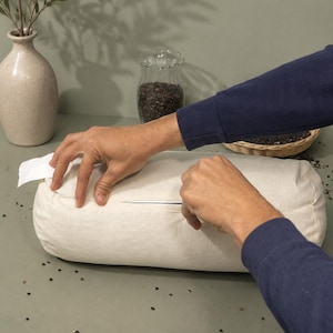 May include: A cylindrical off-white bolster pillow featuring a visible horizontal zipper closure along its side. The pillow is made of a plain, light-colored fabric and is positioned horizontally, showing a clean, simple design without any patterns.
