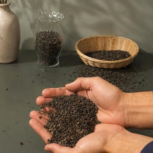 May include: A person's hands cupping a quantity of dark brown buckwheat groats. A wicker basket, a glass jar, and a vase with greenery are in the background. The groats are scattered on a dark green surface.