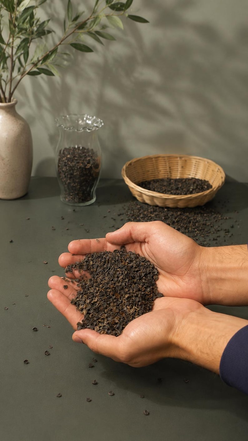 May include: A person's hands cupping a quantity of dark brown buckwheat groats. A wicker basket, a glass jar, and a vase with greenery are in the background. The groats are scattered on a dark green surface.