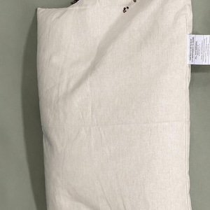 May include: A rectangular off-white fabric pillow case with a zipper closure along the top edge. A pile of small, dark brown buckwheat hulls is visible spilling out from the open zipper. A white fabric tag with black text is attached to the side.
