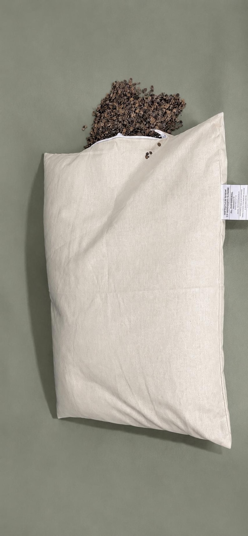 May include: A rectangular off-white fabric pillow case with a zipper closure along the top edge. A pile of small, dark brown buckwheat hulls is visible spilling out from the open zipper. A white fabric tag with black text is attached to the side.