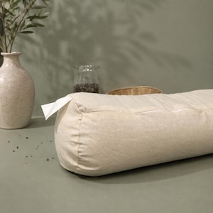 May include: A cylindrical beige bolster cushion with a zip and a white tag. The cushion is made of textured fabric. A vase with olive branches and a glass jar are in the background.