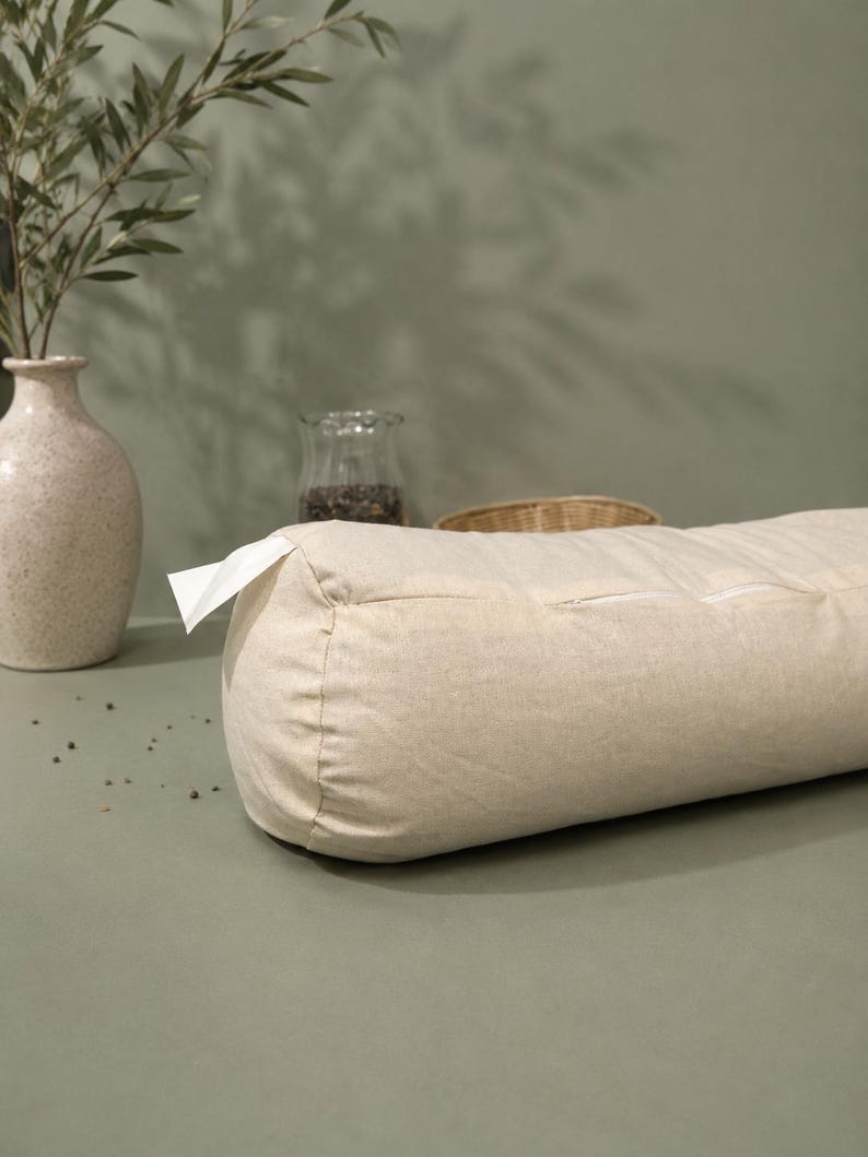 May include: A cylindrical beige bolster cushion with a zip and a white tag. The cushion is made of textured fabric. A vase with olive branches and a glass jar are in the background.