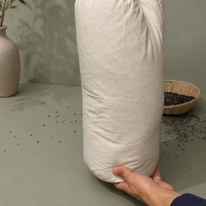 May include: A large, off-white cylindrical bag, held upright. The bag appears to be filled with a granular substance. A small woven basket and a vase with greenery are in the background. The bag is the central focus.
