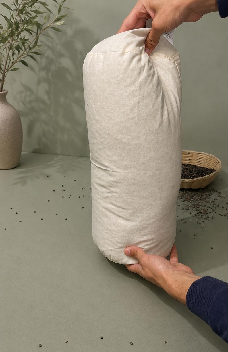 May include: A large, off-white cylindrical bag, held upright. The bag appears to be filled with a granular substance. A small woven basket and a vase with greenery are in the background. The bag is the central focus.