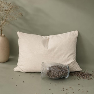 May include: A rectangular beige pillow with a textured fabric cover. In front of the pillow sits a clear glass jar with a ruffled rim, filled with dark brown buckwheat hulls, with some hulls spilled onto the surface in front of the jar.