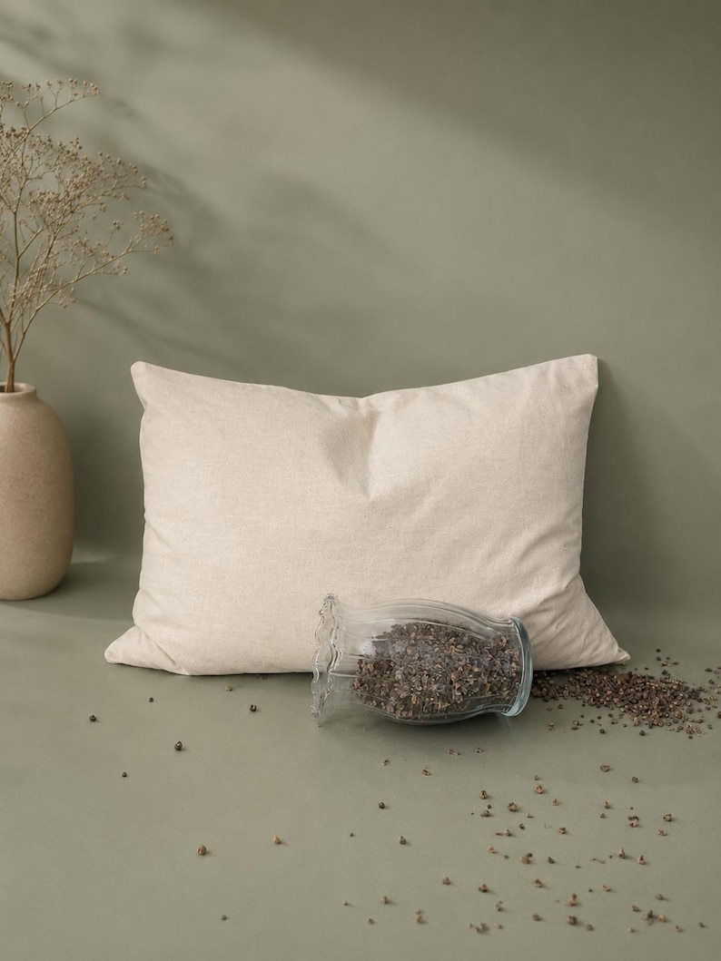 May include: A rectangular beige pillow with a textured fabric cover. In front of the pillow sits a clear glass jar with a ruffled rim, filled with dark brown buckwheat hulls, with some hulls spilled onto the surface in front of the jar.