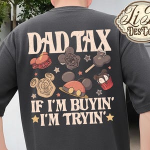 May include: Dark gray t-shirt with the text "DAD TAX" and "IF I'M BUYIN' I'M TRYIN'". The shirt features cartoon graphics of Mickey Mouse, a cupcake, and other treats. The shirt has short sleeves.