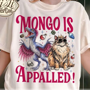 May include: Cream-colored t-shirt featuring a blue dinosaur with pink wings and a cat wearing sunglasses. The text "MONGO IS APPALLED!" is in pink. The shirt also has the text "ORIGINAL DESIGNER".