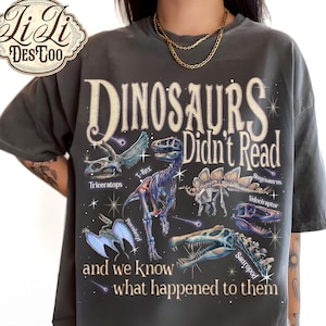 May include: A charcoal gray t-shirt featuring a graphic print of various dinosaur skeletons, including a T-Rex, Triceratops, and Pterodactyl. The text reads: Dinosaurs Didn't Read and we know what happened to them, surrounded by small star illustrations.