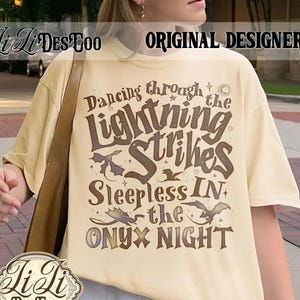 Könnte beinhalten: Cremefarbenes T-Shirt mit dem Text "Dancing through the Lightning Strikes Sleepless IN the Onyx Night" in dunkler brauner Schrift. Das Shirt zeigt auch Drachenillustrationen. Der Markenname "Li Li Des Too" ist sichtbar.