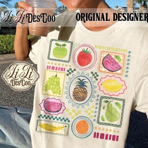 May include: Cream-colored t-shirt featuring a colorful fruit-themed design. The design includes illustrations of various fruits like apples, strawberries, watermelons, grapes, pineapples, lemons, pears, bananas, and oranges, all within decorative frames. The text "ORIGINAL DESIGNER" is visible.