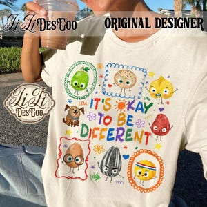 Könnte beinhalten: Cremefarbenes T-Shirt mit einem farbenfrohen Aufdruck mit dem Text "IT'S OKAY TO BE DIFFERENT". Das Design zeigt Cartoon-Essensfiguren und einen Hund, alle in dekorativen Rahmen. Ein lässiges Kleidungsstück.