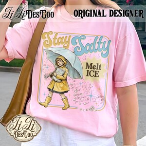 Pode incluir: Camiseta rosa claro com um gr&aacute;fico de uma menina de capa de chuva e botas amarelas segurando um guarda-chuva. O texto "Stay Salty" e "Melt ICE" tamb&eacute;m est&atilde;o na camiseta. As palavras "Original Designer" s&atilde;o impressas no topo.