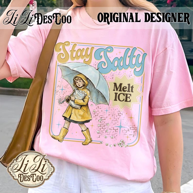 Pode incluir: Camiseta rosa claro com um gr&aacute;fico de uma menina de capa de chuva e botas amarelas segurando um guarda-chuva. O texto "Stay Salty" e "Melt ICE" tamb&eacute;m est&atilde;o na camiseta. As palavras "Original Designer" s&atilde;o impressas no topo.