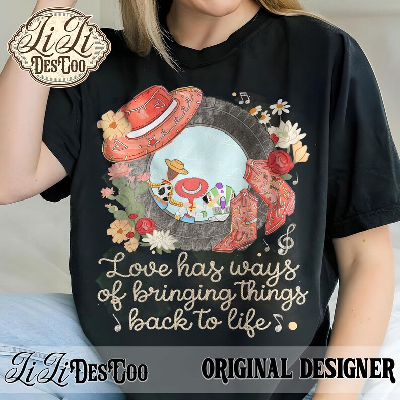 Puede incluir: Una camiseta negra con un estampado gr&aacute;fico de un marco de neum&aacute;tico que contiene personajes de dibujos animados, un sombrero de vaquero rojo y botas rojas. El dise&ntilde;o incluye detalles florales y el texto, Love has ways of bringing things back to life.